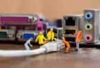 Des figurines miniatures de travailleurs installent un câble Ethernet RJ45 devant une carte mère d'ordinateur, illustrant la comparaison technique entre les câbles Cat5e et Cat6.