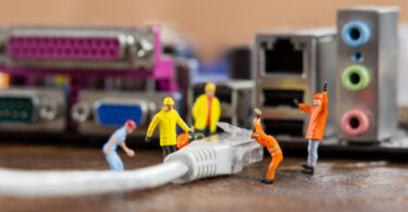 Des figurines miniatures de travailleurs installent un câble Ethernet RJ45 devant une carte mère d'ordinateur, illustrant la comparaison technique entre les câbles Cat5e et Cat6.