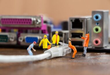 Des figurines miniatures de travailleurs installent un câble Ethernet RJ45 devant une carte mère d'ordinateur, illustrant la comparaison technique entre les câbles Cat5e et Cat6.