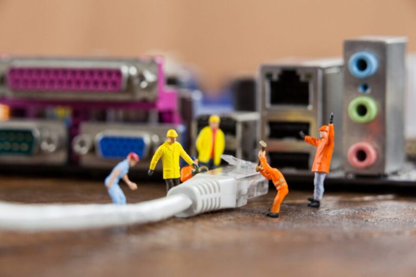 Des figurines miniatures de travailleurs installent un câble Ethernet RJ45 devant une carte mère d'ordinateur, illustrant la comparaison technique entre les câbles Cat5e et Cat6.
